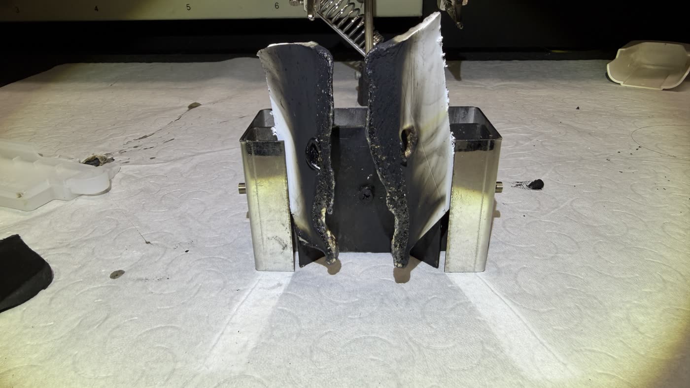 Burnt material sample in the holder after the test, heavily charred and deformed.